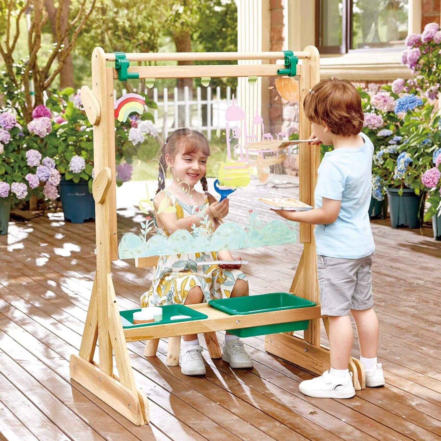 Two children playing with Hape Outdoor Art Easel outdoors on a wooden deck.
