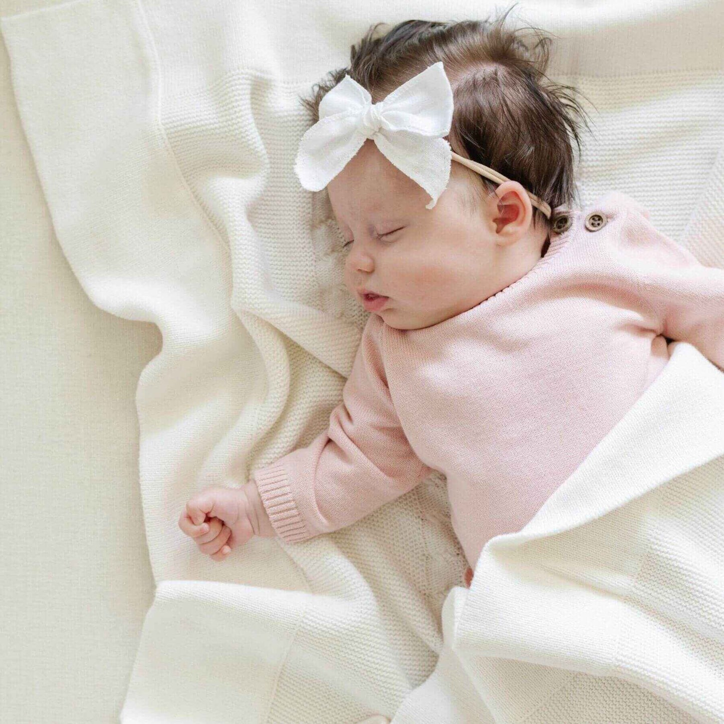 Baby in pink outfit with white bow lying on Saranoni Heirloom Cotton Knit Receiving Blanket Cream