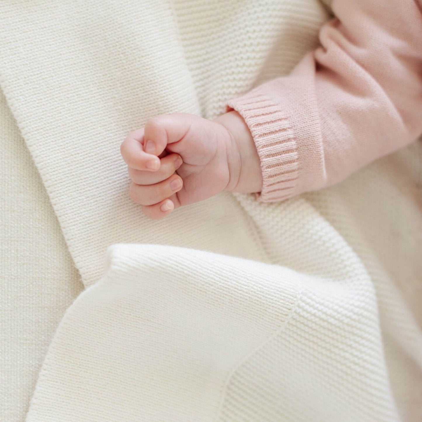 Close-up of a baby's hand in a pink sweater on Saranoni Heirloom Cotton Knit Receiving Blanket Cream