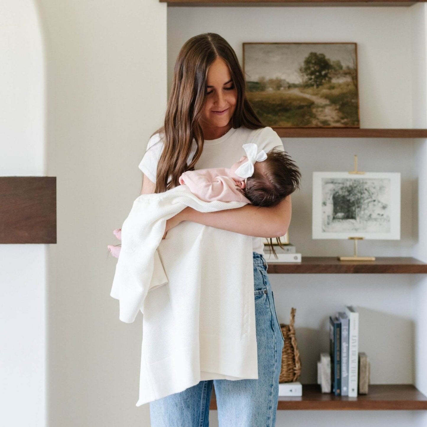 Woman holding a baby with Saranoni Heirloom Cotton Knit Receiving Blanket in a home setting with shelves in the background