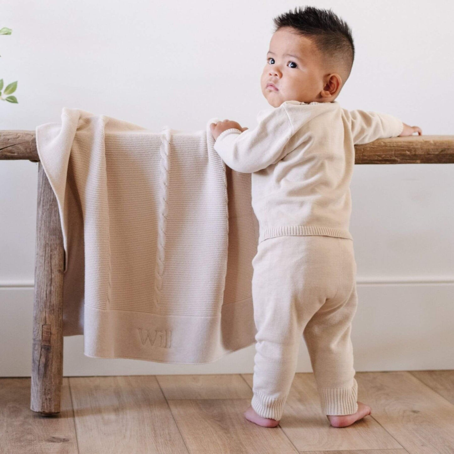 Child in a beige outfit standing next to a wooden bench with Saranoni Heirloom Cotton Knit Receiving Blanket