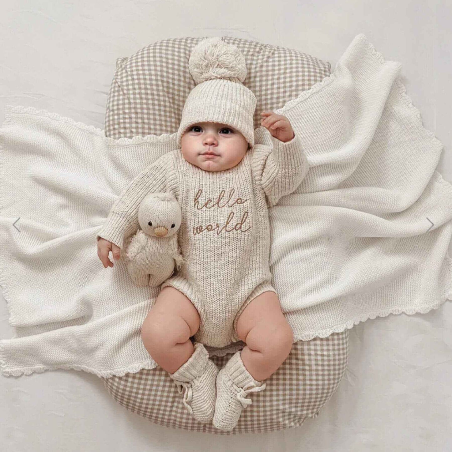 Baby in Newborn Hello World Knitted Bodysuit Set, lying on a checkered cushion with a white blanket.