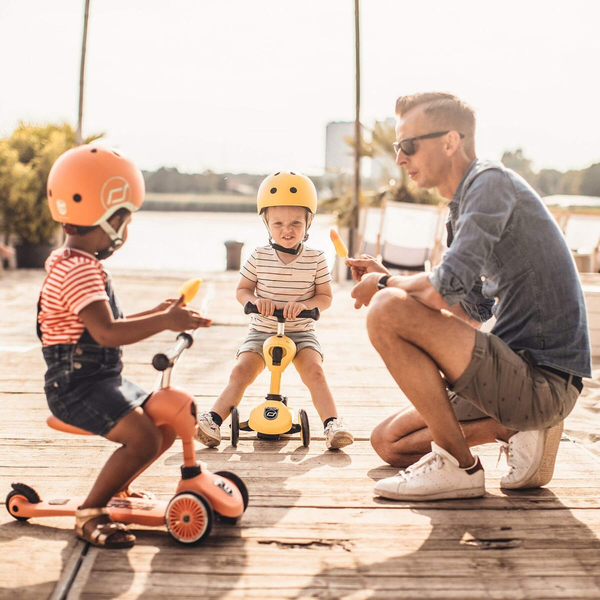 Two children on scooters with a man sitting nearby on a wooden dock.