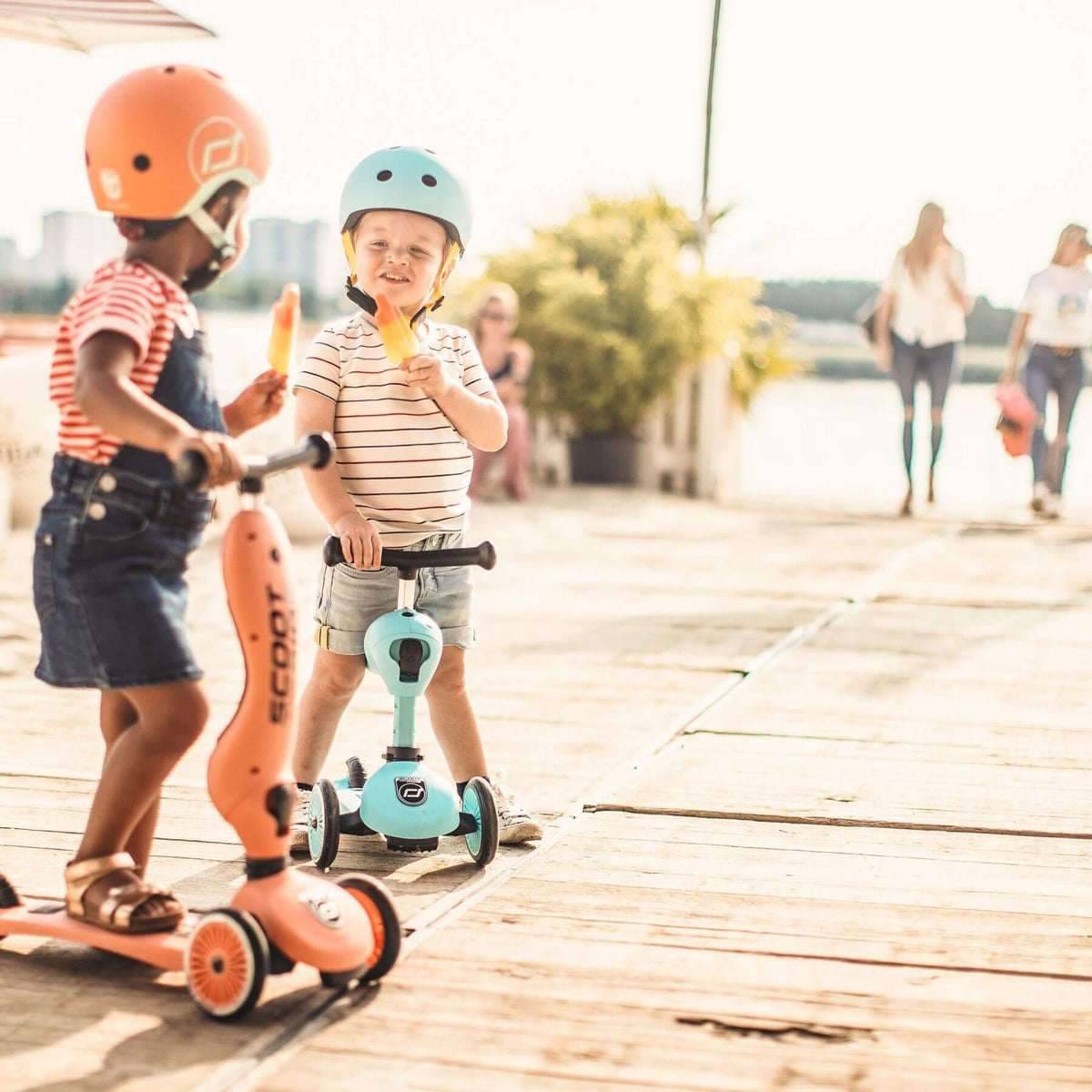 Two children on scooters with helmets at a waterfront