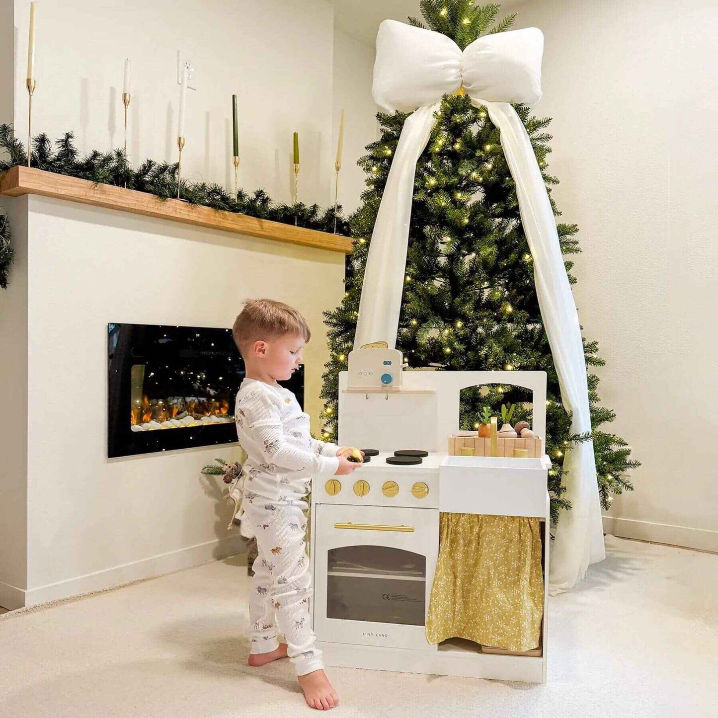 Child playing with Tiny Land® Honey Kitchenette - Little Chef's First Kitchen in a room with a Christmas tree and fireplace.