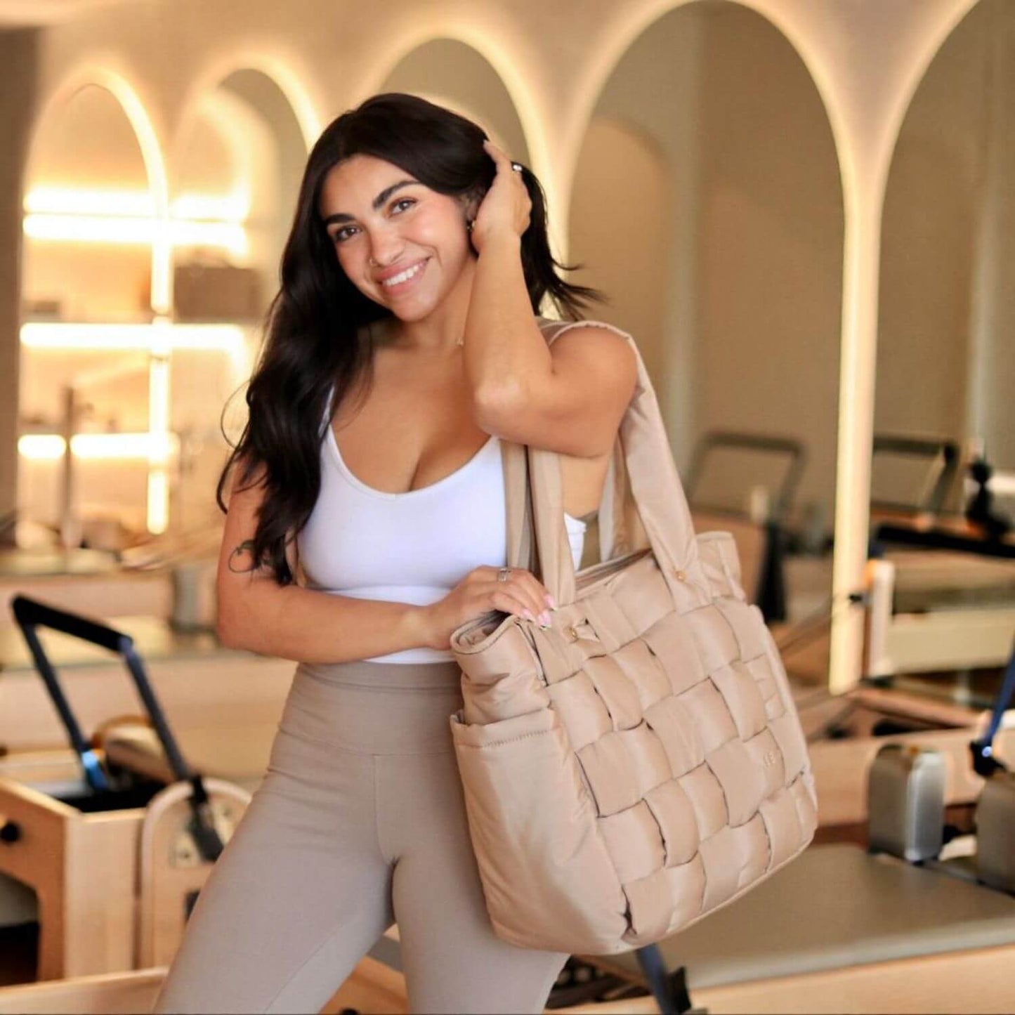 A woman in a white tank top and beige leggings holds Luli Bebé Infinite Diaper Bag - Latte in a bright, modern studio with arched windows in the background.