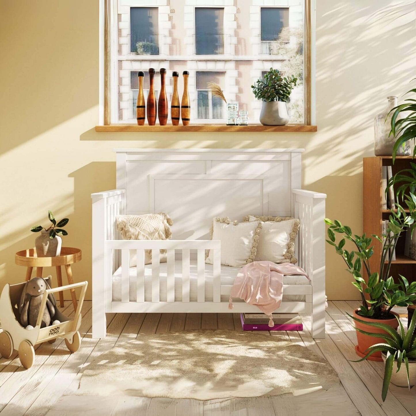 Milk Street Baby "Just White" OG 4-in-1 Convertible Crib with pillows in a sunlit nursery with plants and toys.