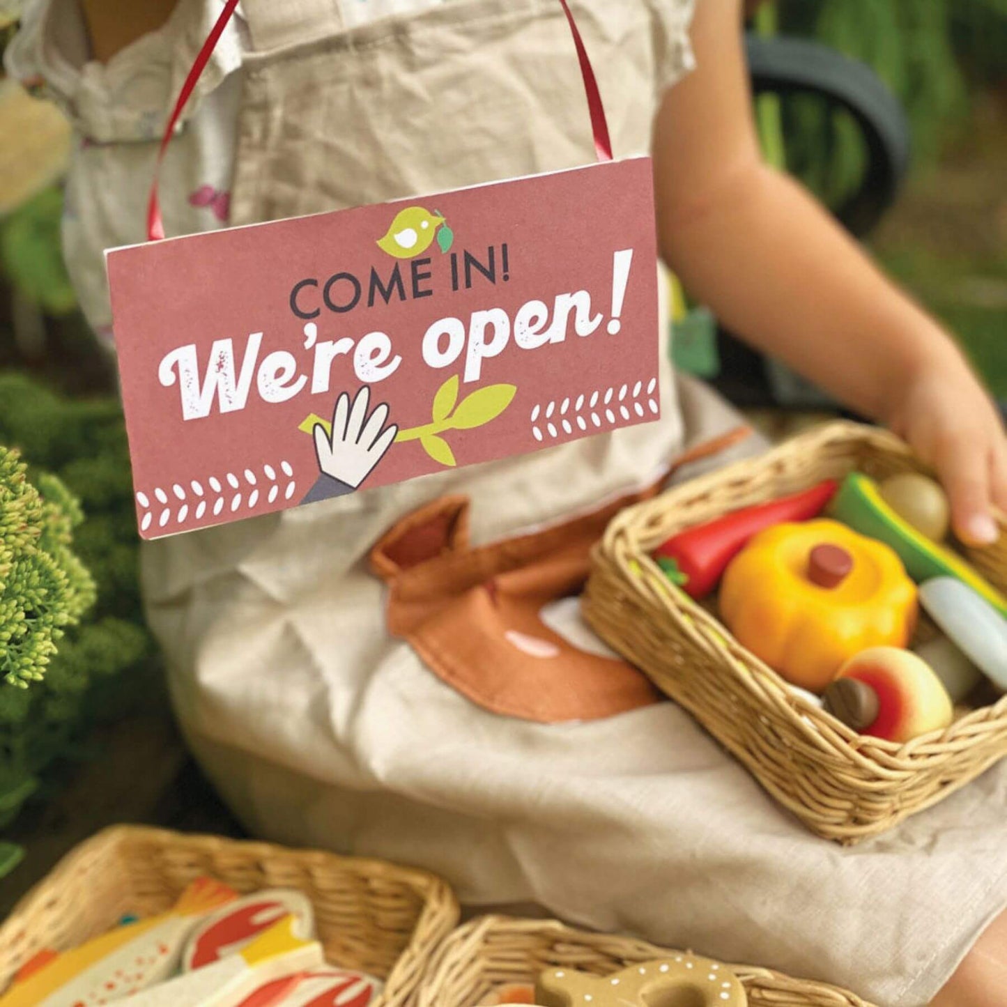Person holding a basket of toy vegetables with a 'Come in! We're open!' sign.