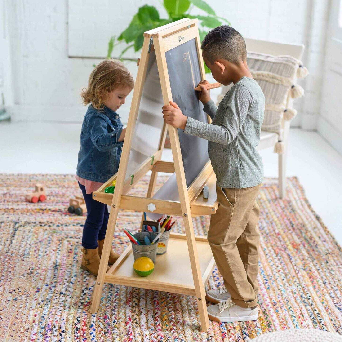 Two children using a Little Partners Folding Artist EZ Easel Natural with paper and art supplies in a bright room.