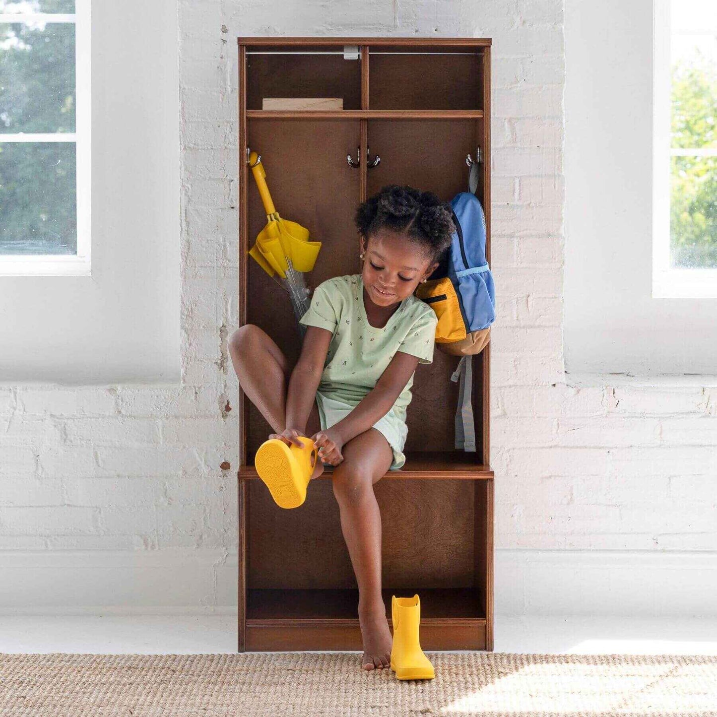 Child sitting on Little Partners Learn 'N Store Cubby with shoes and backpacks, in a room with white brick walls.