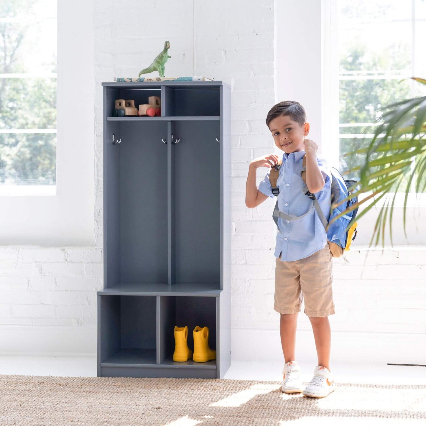 Child with a backpack standing next to a Little Partners Learn 'N Store Cubby Grey with shoes and toys inside.