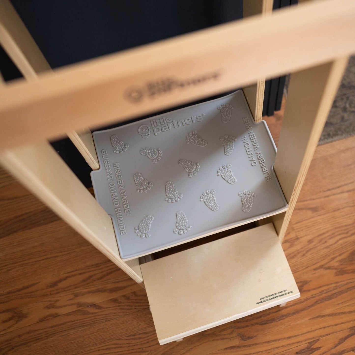 Wooden shelf with a Little Partners Silicone Mat featuring footprints and text, on a wooden floor.