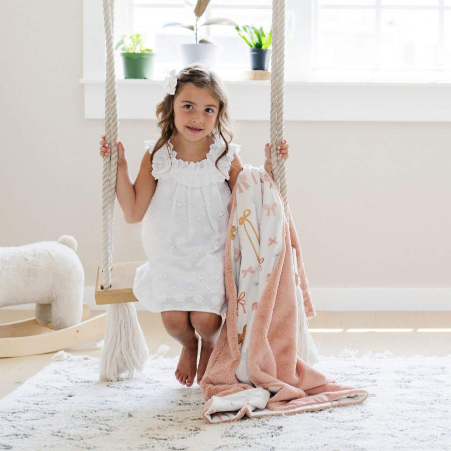 Young girl in a white dress sitting on a wooden swing with Saranoni Minky/Lush Toddler Blanket draped over her, in a bright room with plants.
