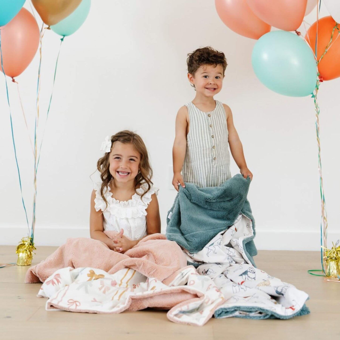 Two children surrounded by colorful balloons and Saranoni Minky/Lush Toddler Blankets on a light background