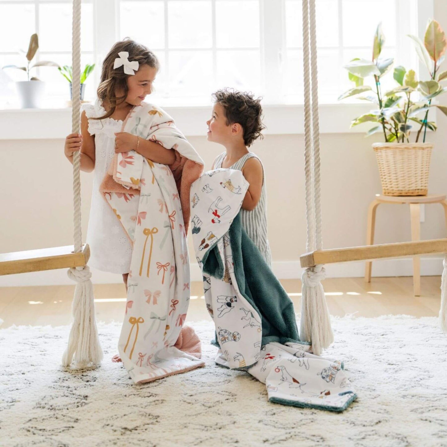 Two children holdingSaranoni Minky/Lush Toddler Blankets on a swing in a bright room with plants.