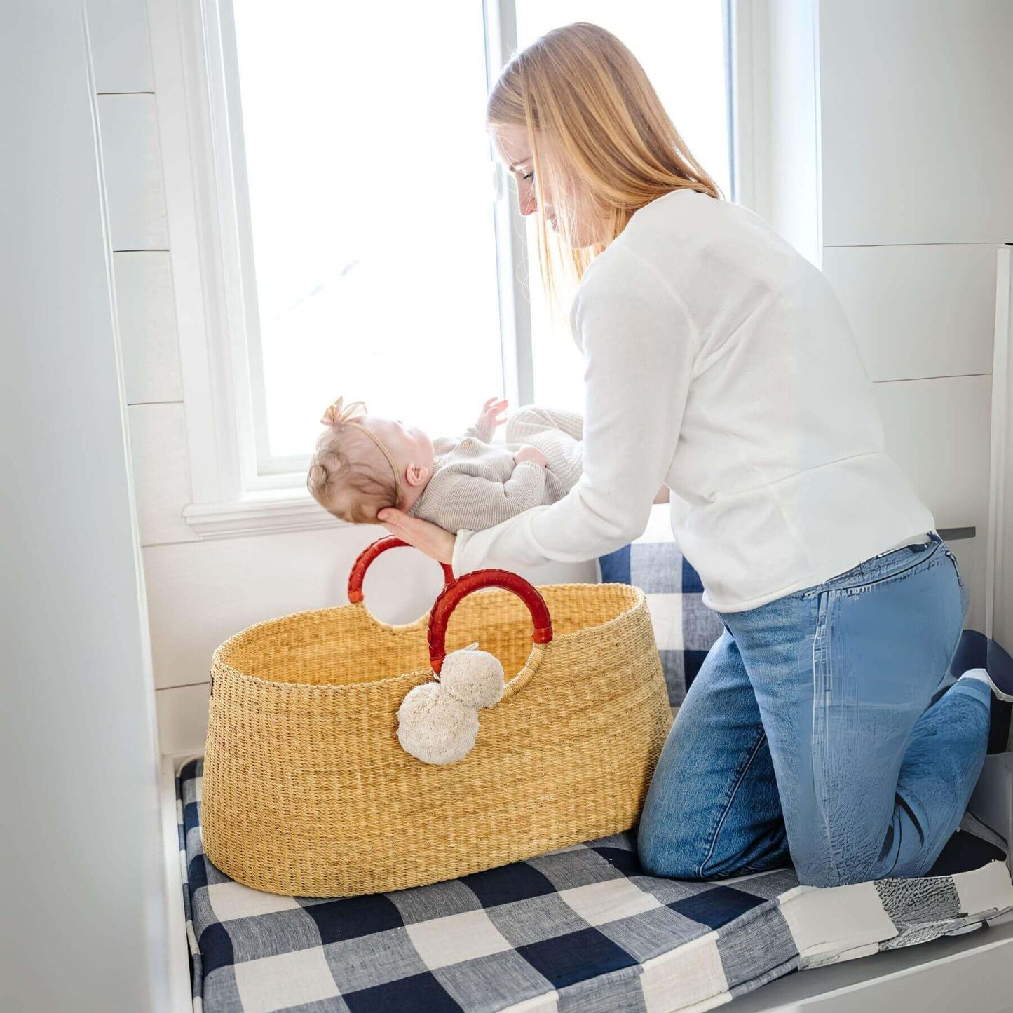 Woman kneeling next to African Moses Basket Cognac Circle Handle with a baby inside, in a home setting.