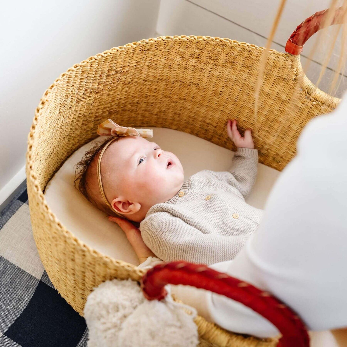 Baby lying in African Moses Basket Cognac Circle Handle with a soft toy, surrounded by plaid bedding.
