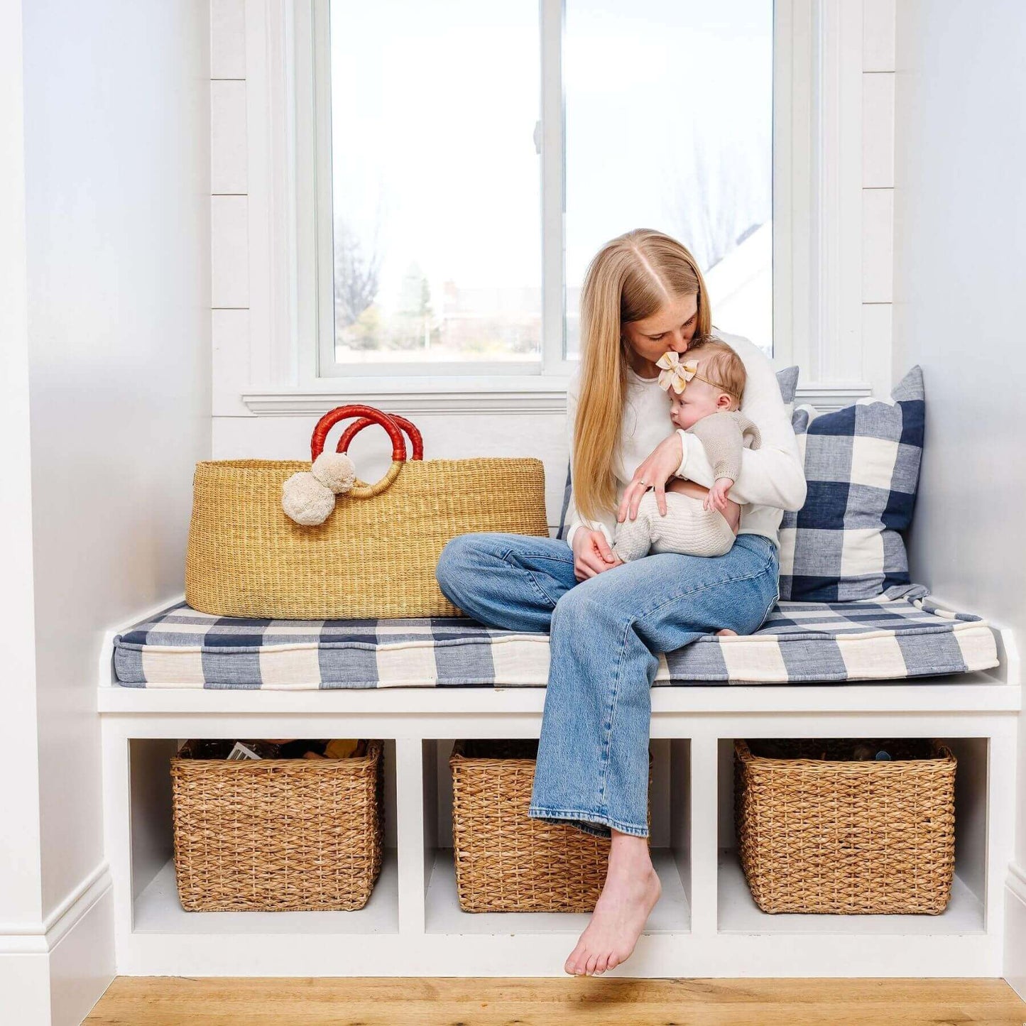 Woman sitting on a bench with a child holding a teddy bear, surrounded by African Moses Basket Cognac Circle Handle and a plaid cushion.