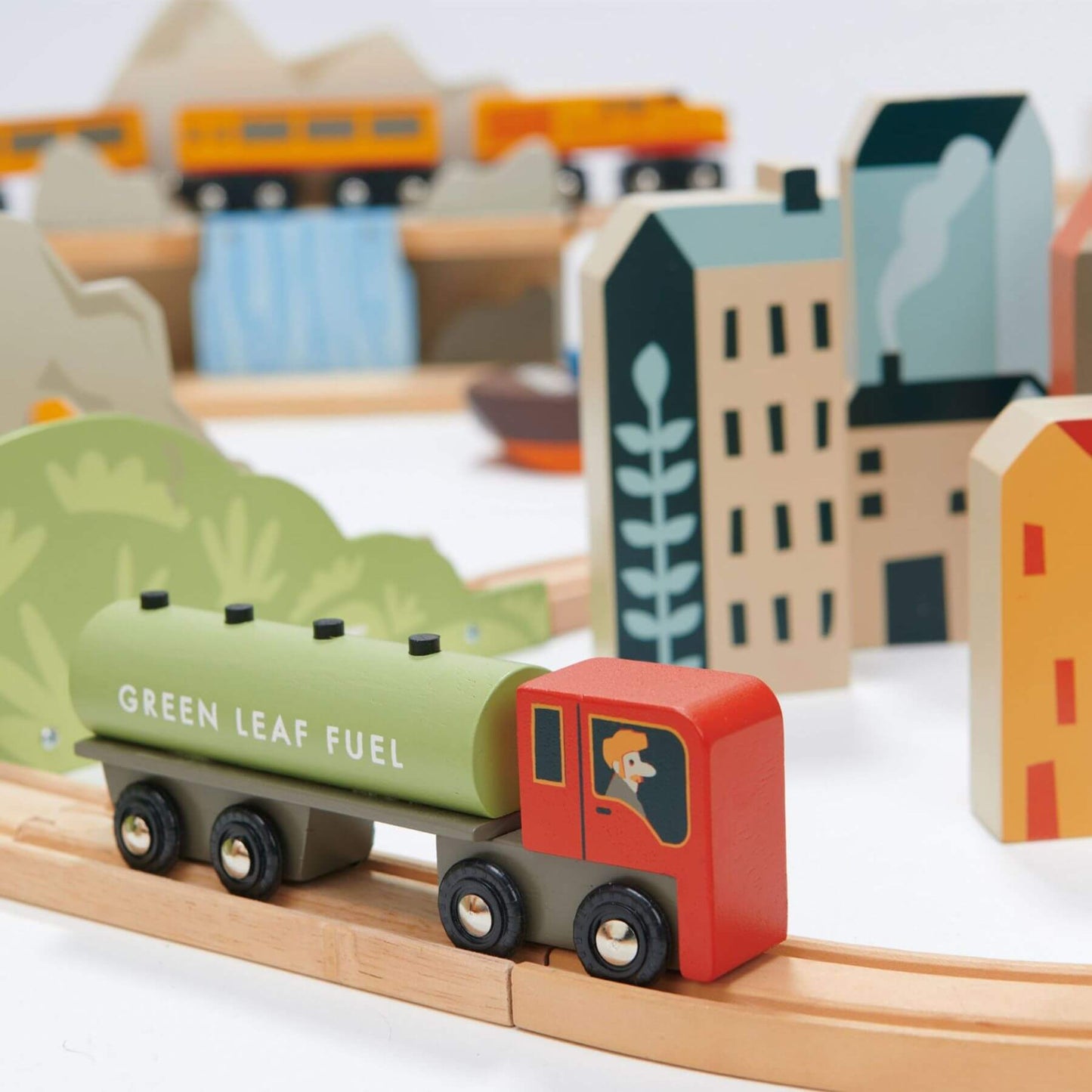 Wooden toy train set with a green leaf fuel car on a track, surrounded by small buildings and trees.
