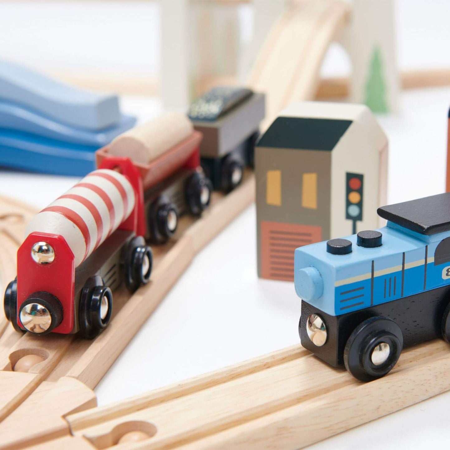 Wooden toy train set on tracks with a red and white striped engine and blue and black car, alongside small buildings and traffic lights.