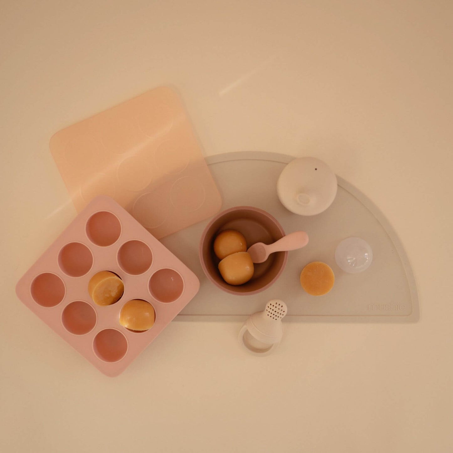 Mushie Baby Food Freezer Tray with yellow balls, brown bowl with spoon, and small containers on a light surface.
