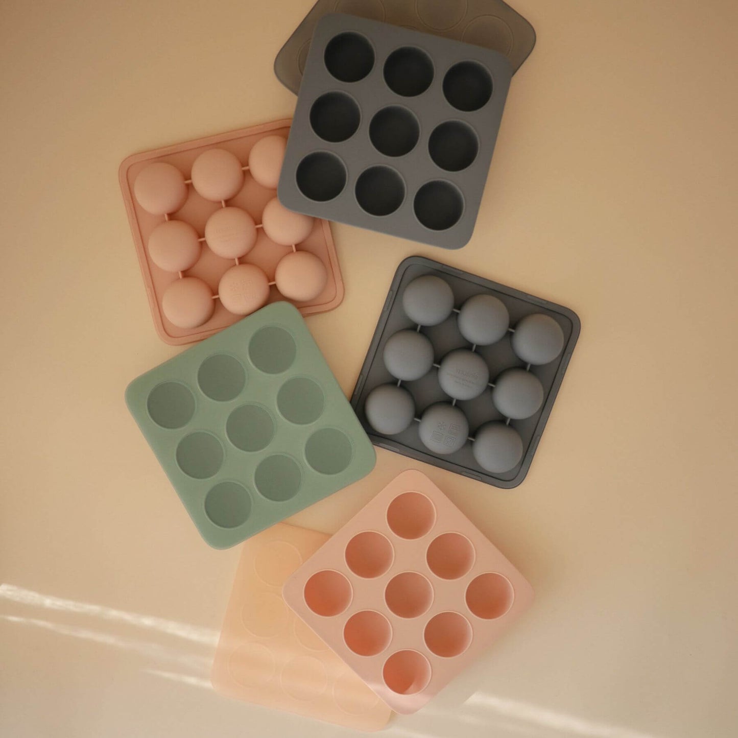 Collection of Mushie Baby Food Freezer Trays in various colors on a beige surface