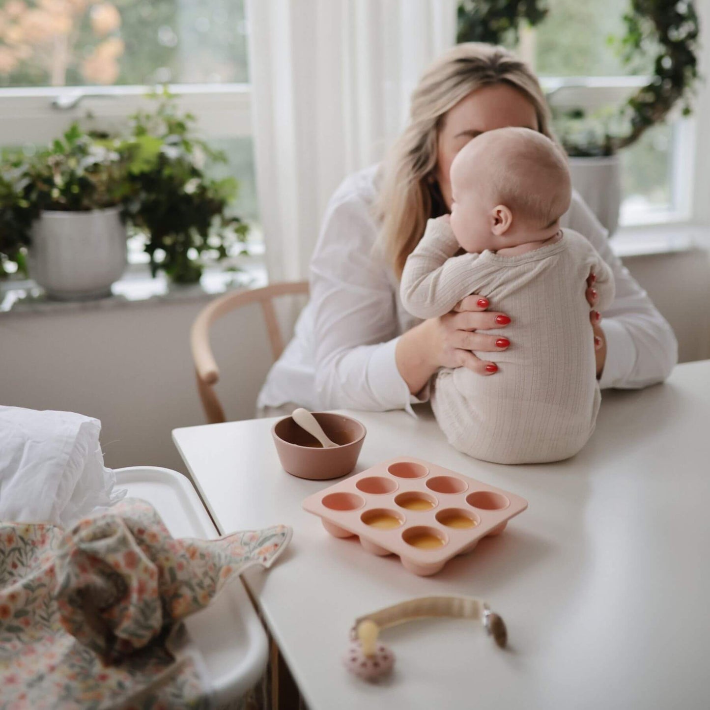 Woman holding a baby in a home setting with a table and various items.