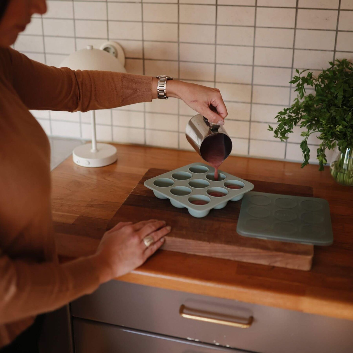 Person pouring liquid into a Mushie Baby Food Freezer Tray Cambridge Blue on a wooden cutting board in a kitchen.