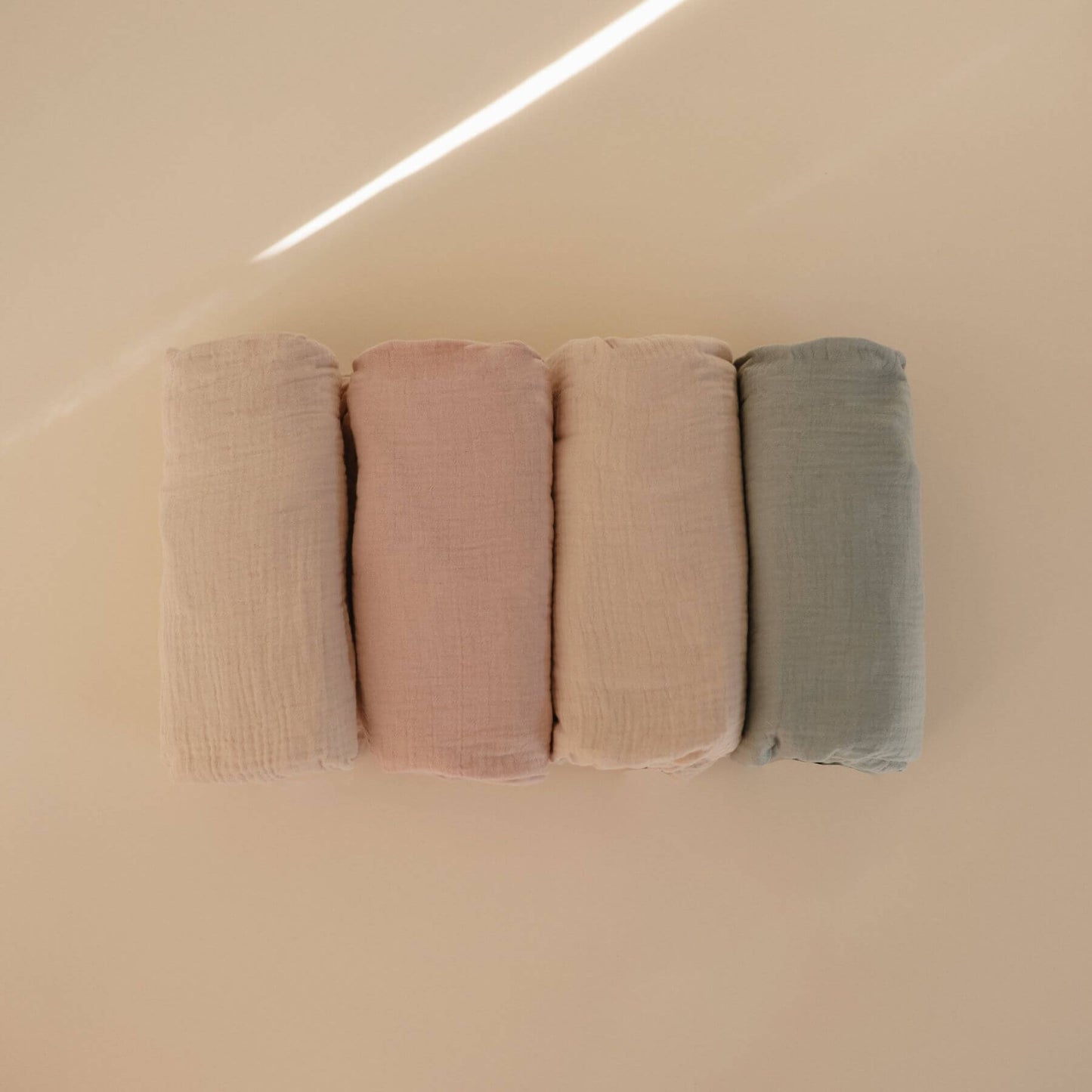 Four mushie muslin crib sheets in different colors arranged on a beige surface.