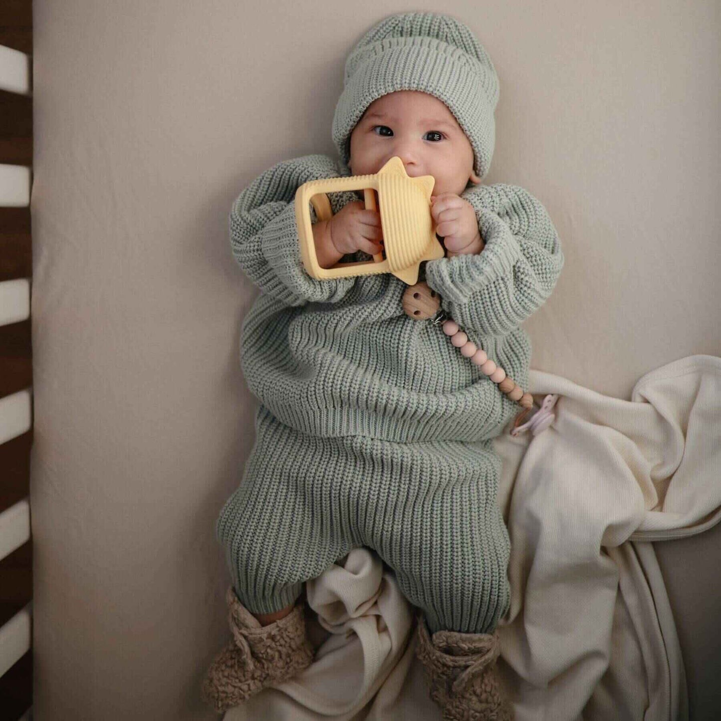 Baby wrapped in a green knitted outfit holding a yellow toy against a neutral background