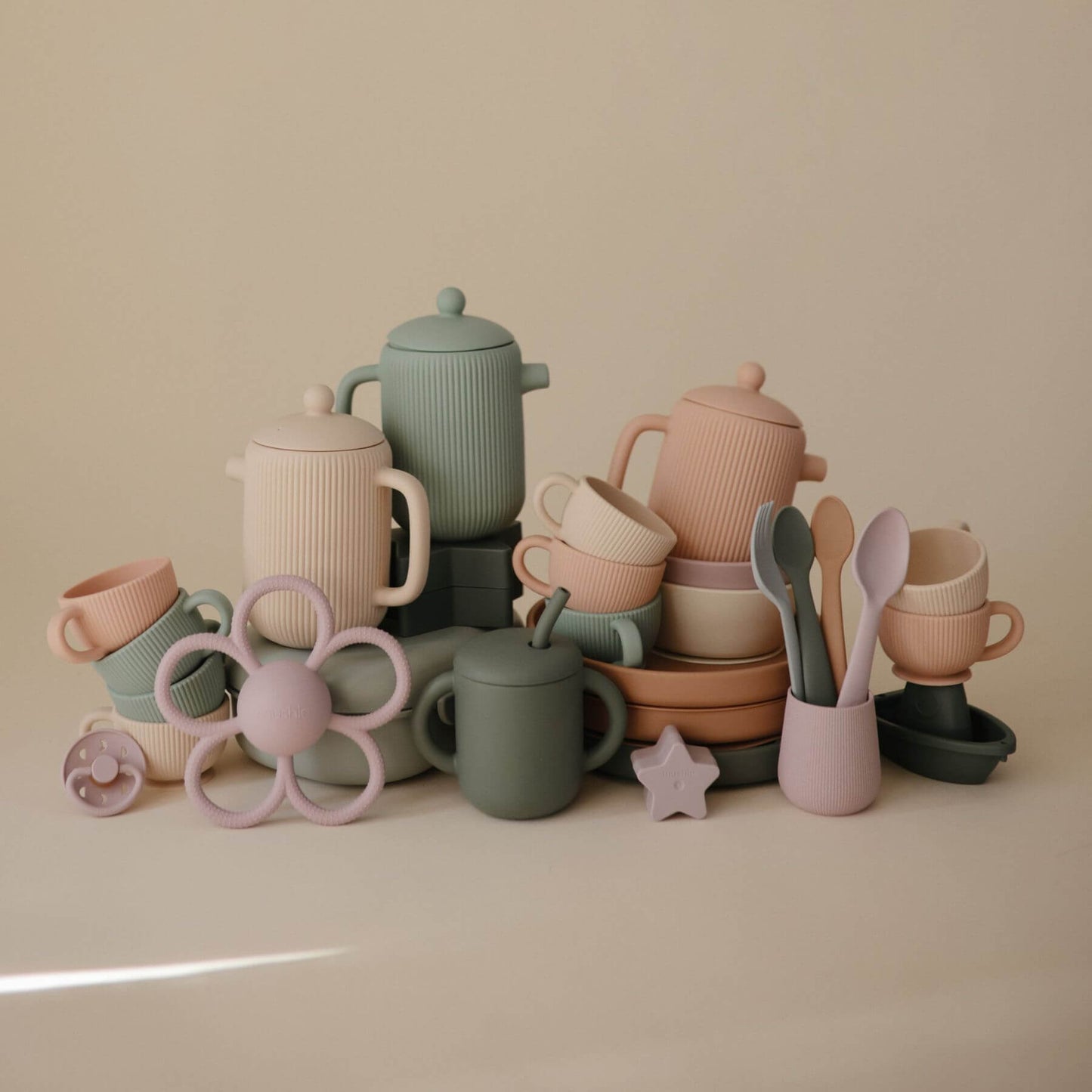 Collection of Mushie Silicone Tea Play Set and toys on a beige surface.