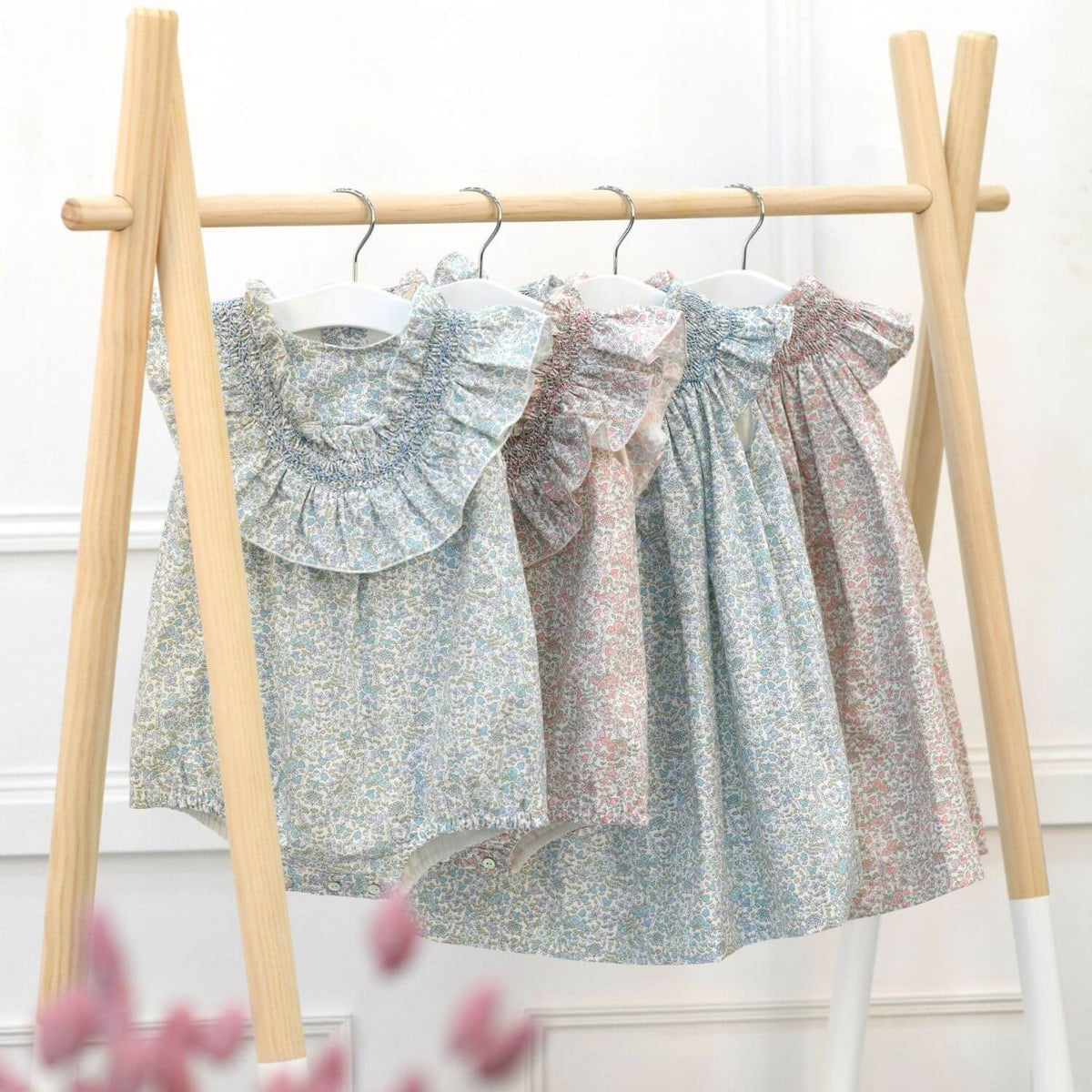 Three children's sleeveless dresses with ruffled collars and flared skirts hanging on a wooden A-frame rack