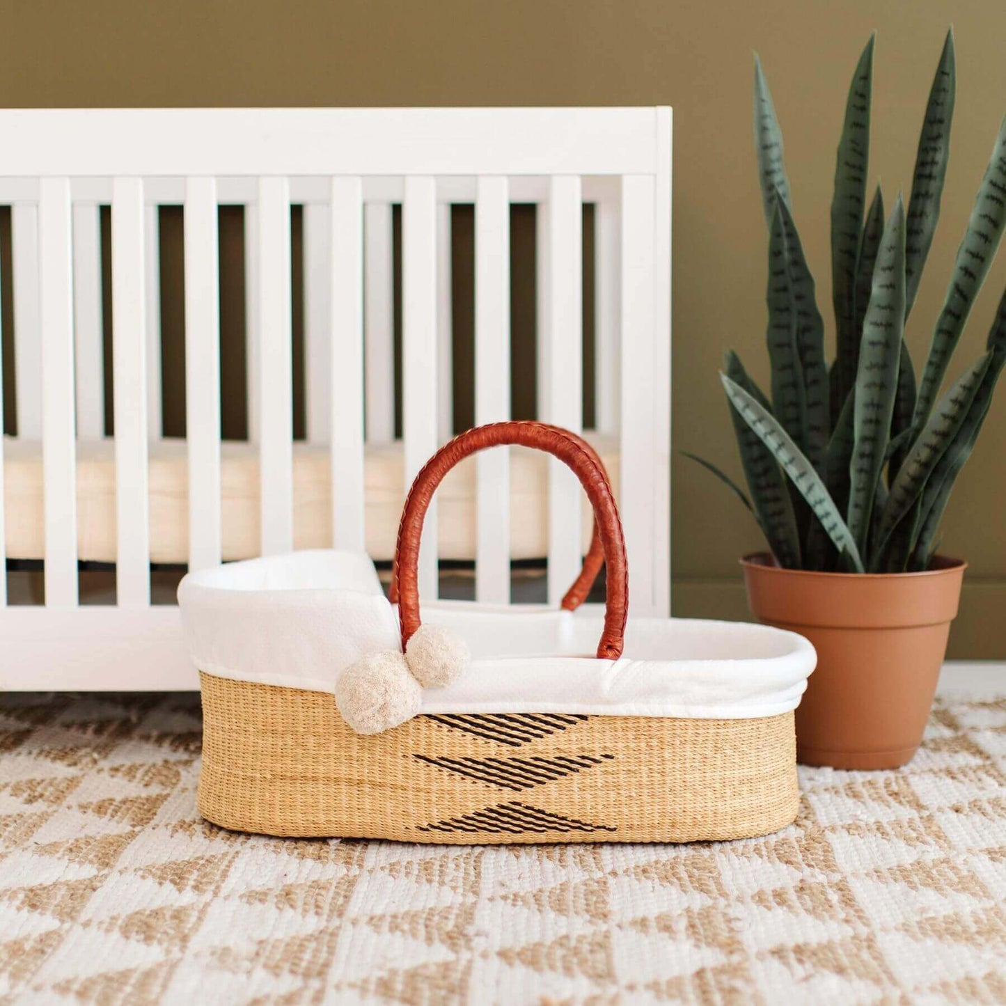 Plum+Sparrow Oak African Moses Basket in front of a white crib with a plant in the background.