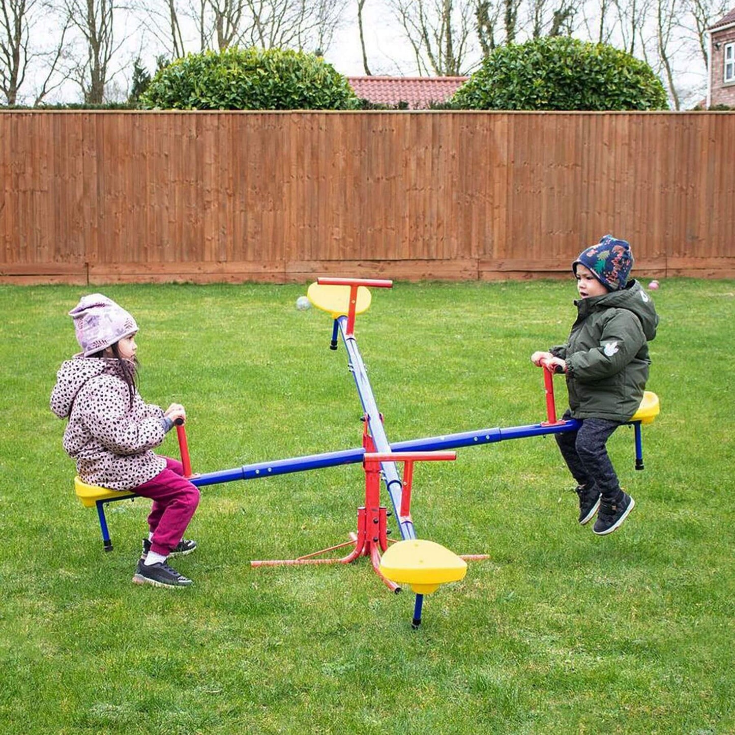Two children playing on a seesaw in a backyard with a wooden fence and trees in the background.