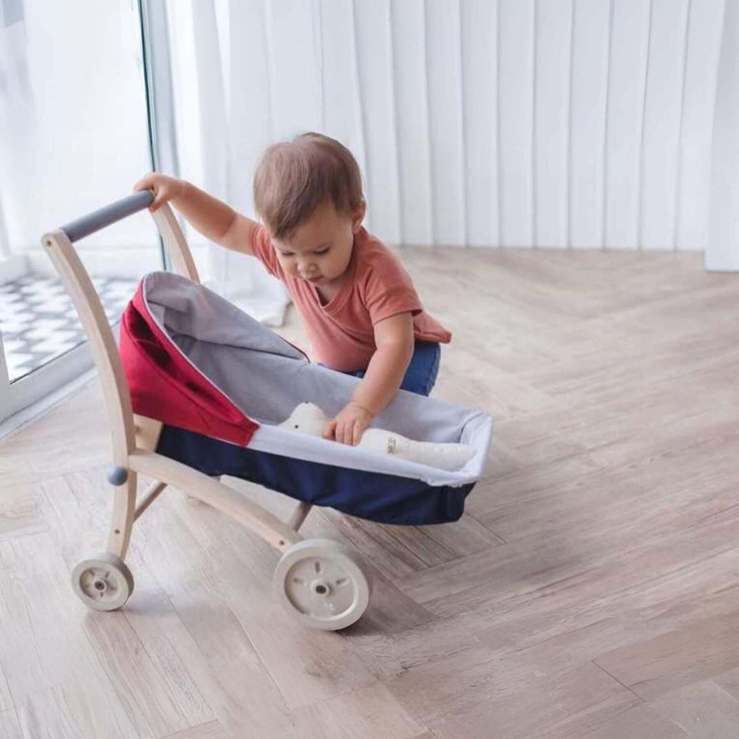 Child playing with PlanToys Doll Stroller indoors on a wooden floor.