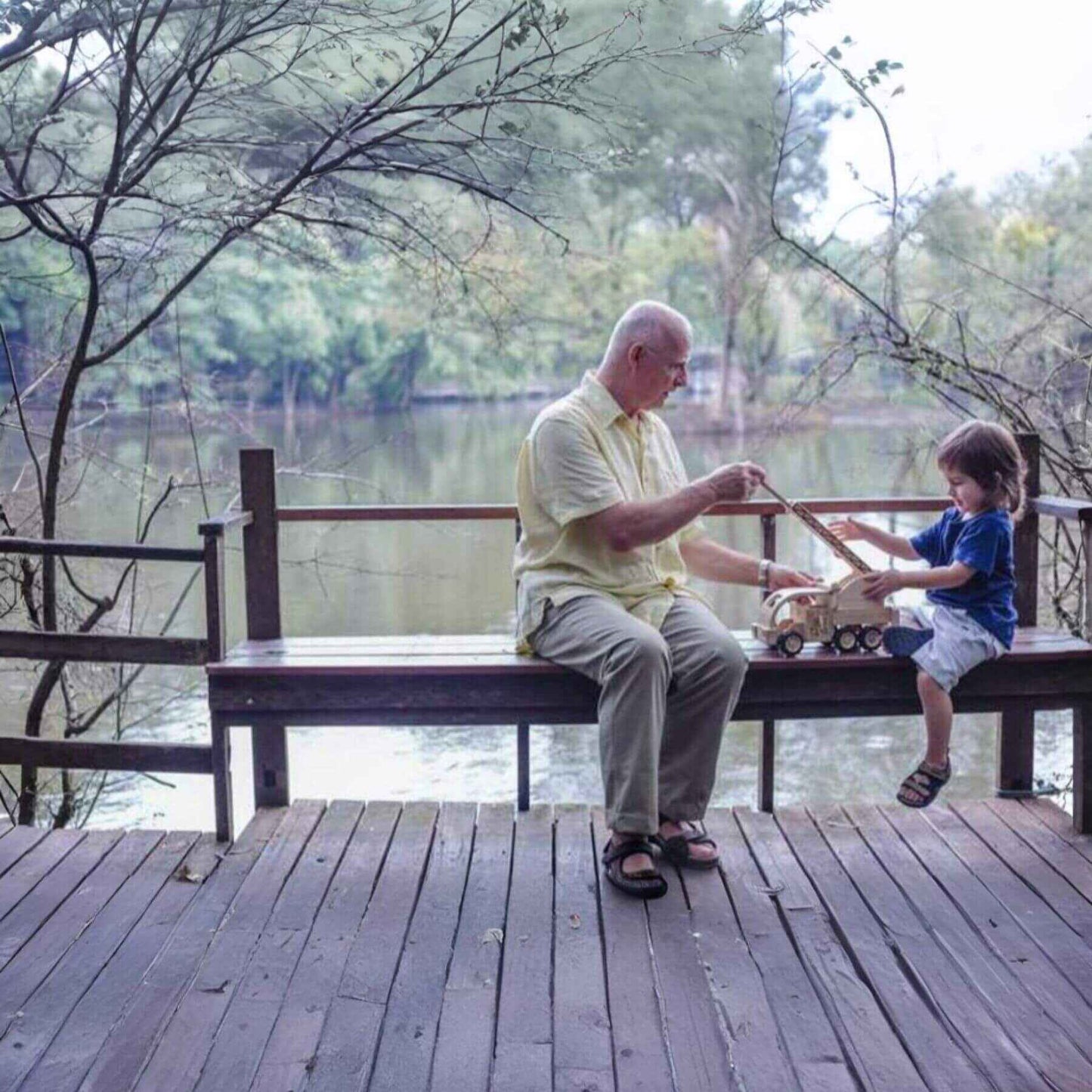 Man and child sitting on a wooden bench by a lake, playing with PlanToys Fire Truck