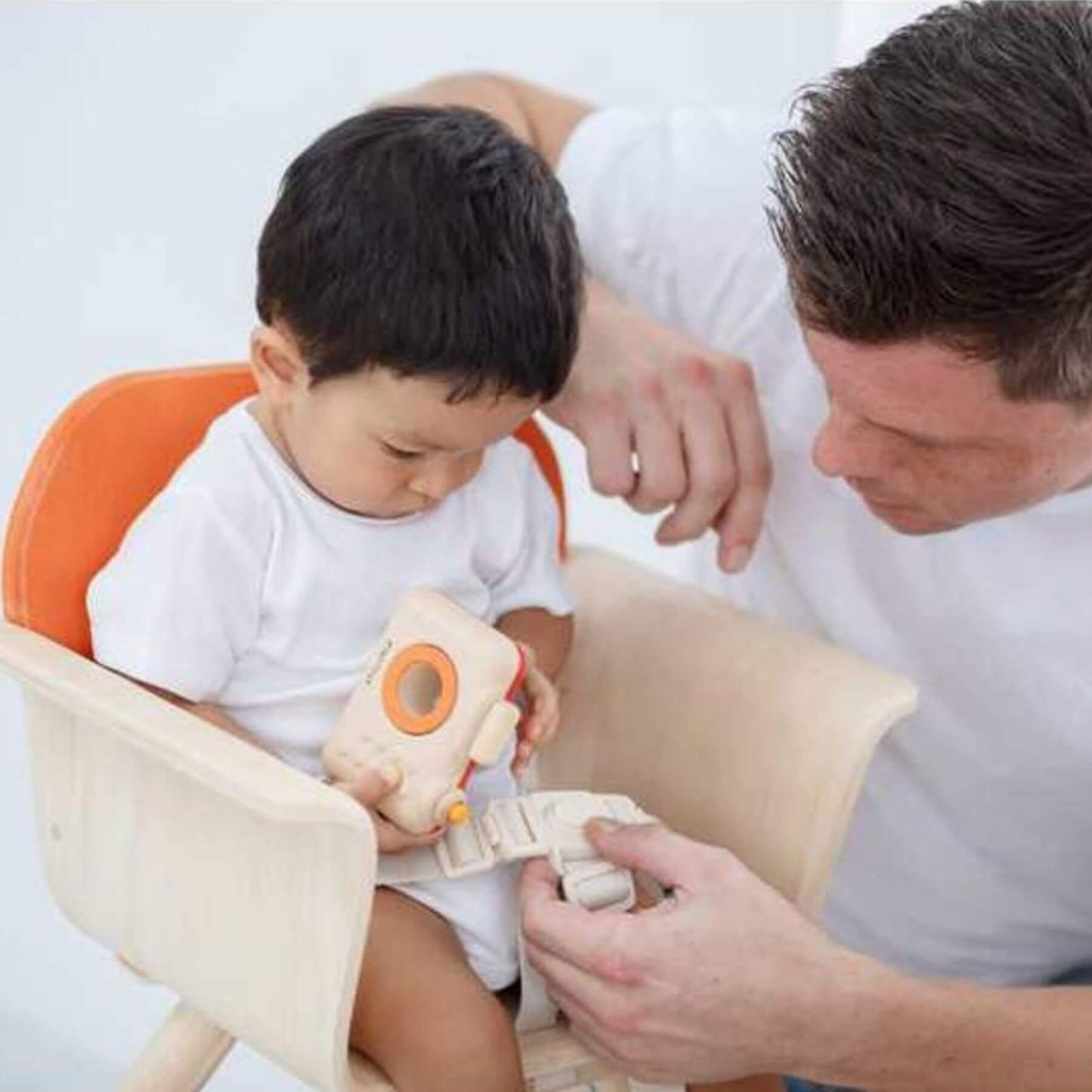 Child sitting in PlanToys High Chair - Orange with a toy, being assisted by an adult.