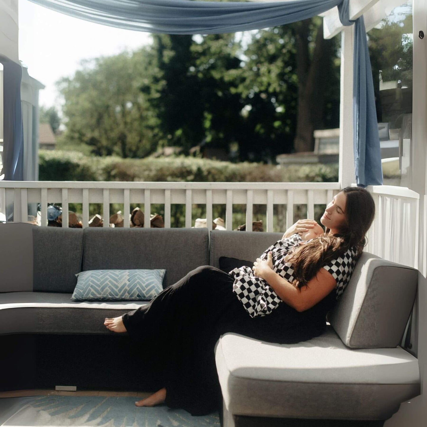 Woman lying on a gray outdoor sofa, holding a baby in Little Mae & Co Race Ring Sling Carrier with a scenic background