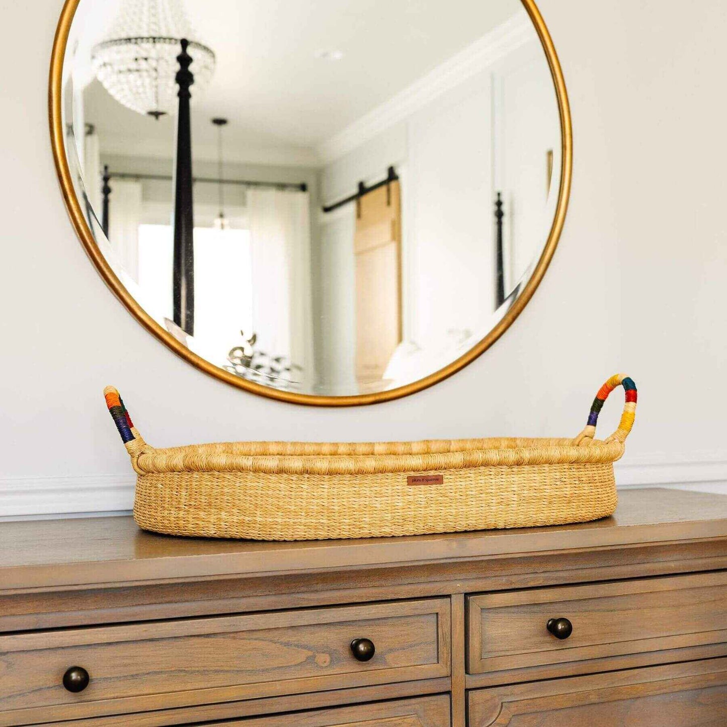Plum+Sparrow Rainbow Handle Changing Basket on a wooden dresser below a round mirror.
