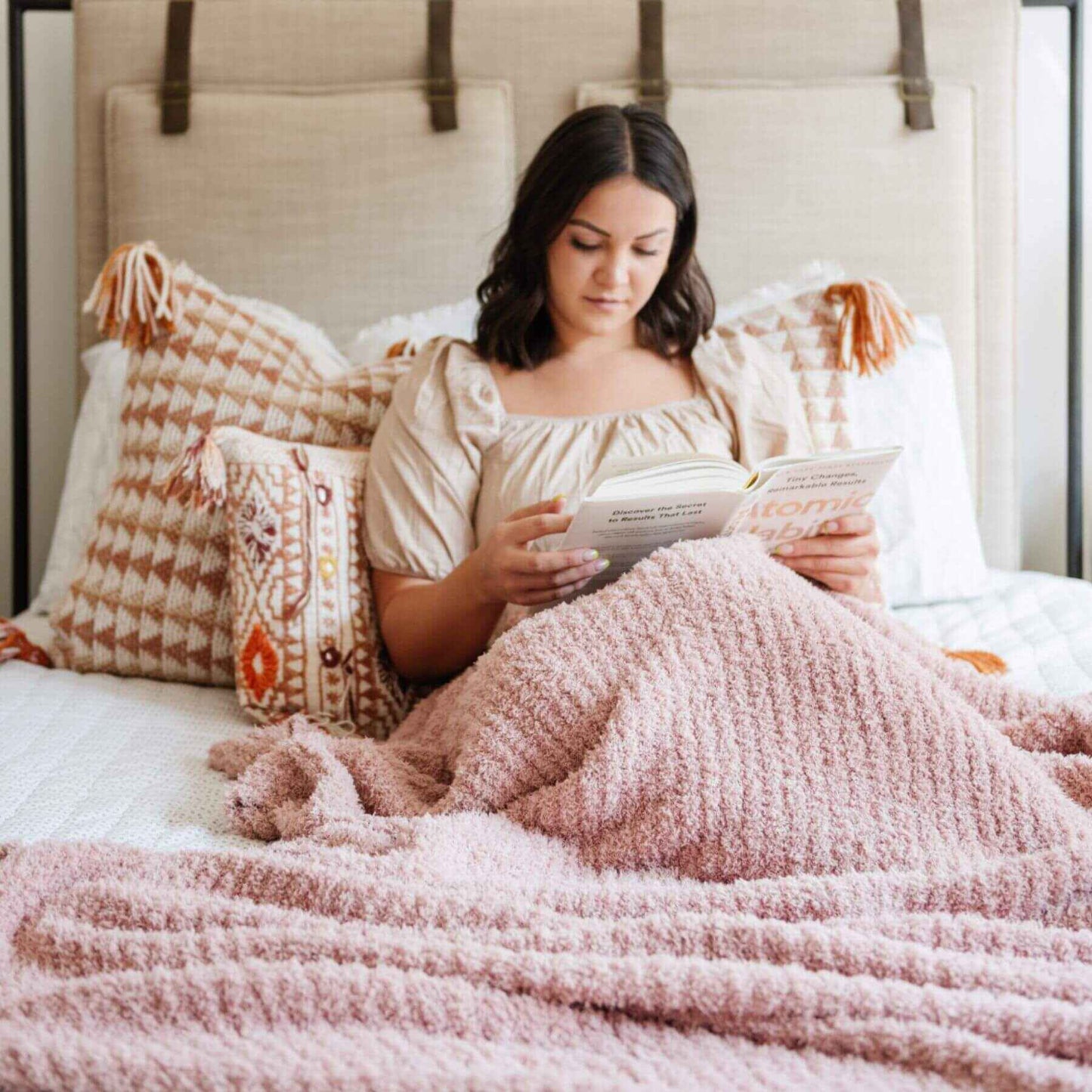 Woman reading a book on a bed with Ribbed Bamboni® Throw Blankets Cameo and patterned pillows.