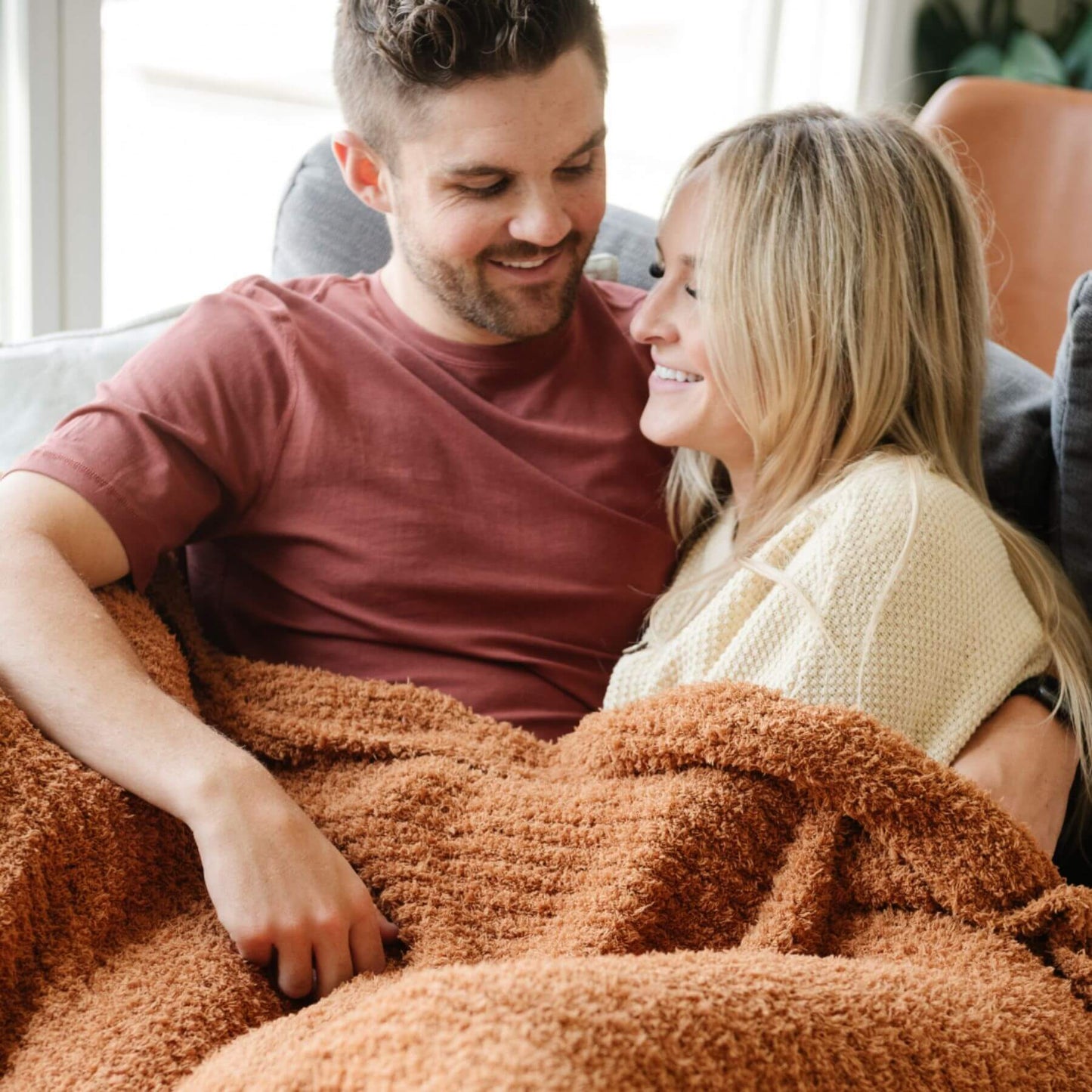 Man and woman sitting together under Ribbed Bamboni® Throw Blankets Ginger, smiling and looking at each other.