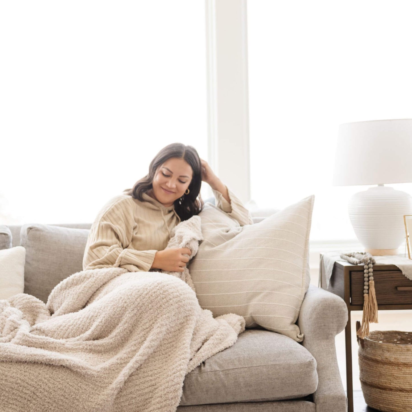 Woman sitting on a couch wrapped in Ribbed Bamboni® Throw Blankets Moonbeam and pillow in a cozy living room.