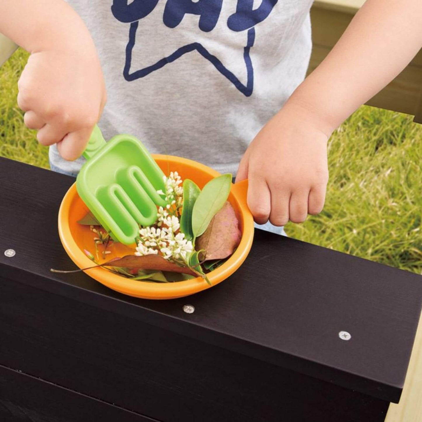 Child playing with a green spatula and orange bowl on a black surface outdoors.