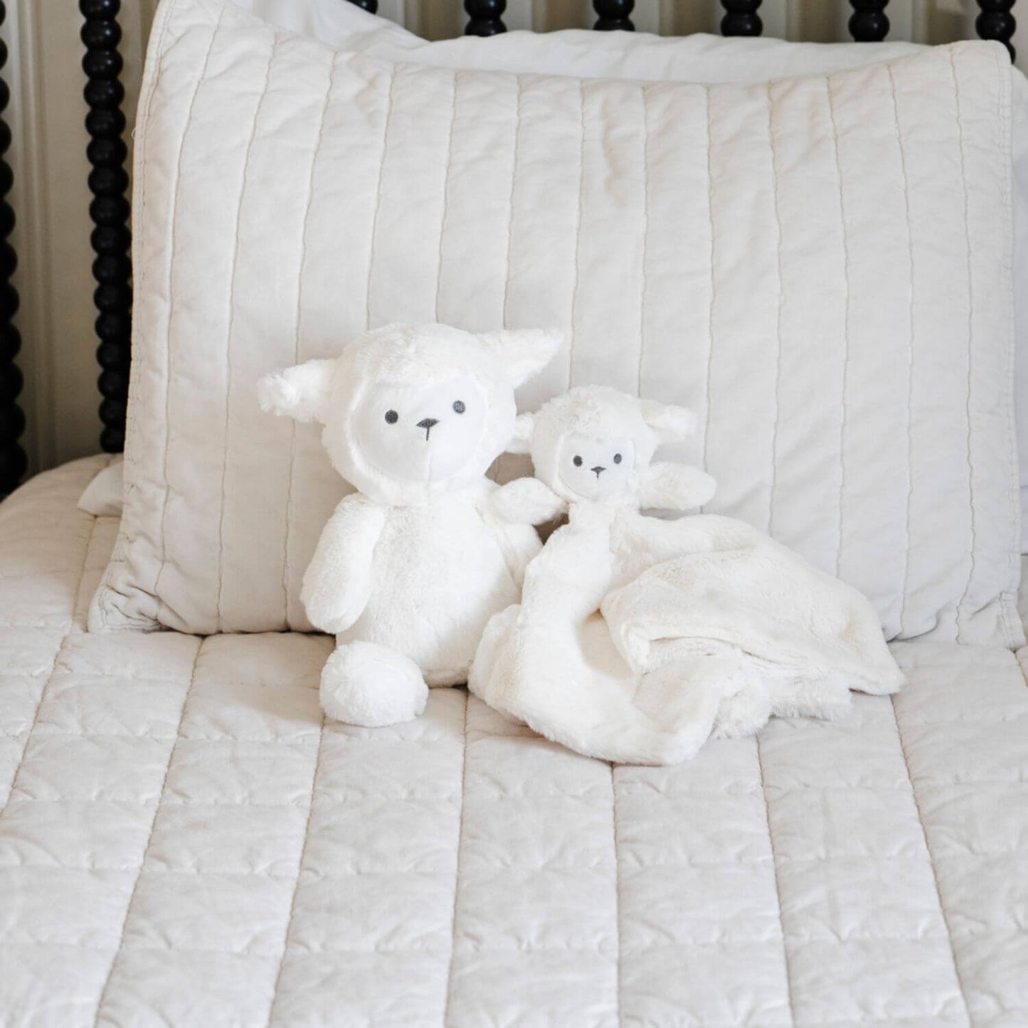 Two Saranoni Stuffed Animal lambs on a striped bedspread with a pillow in the background.