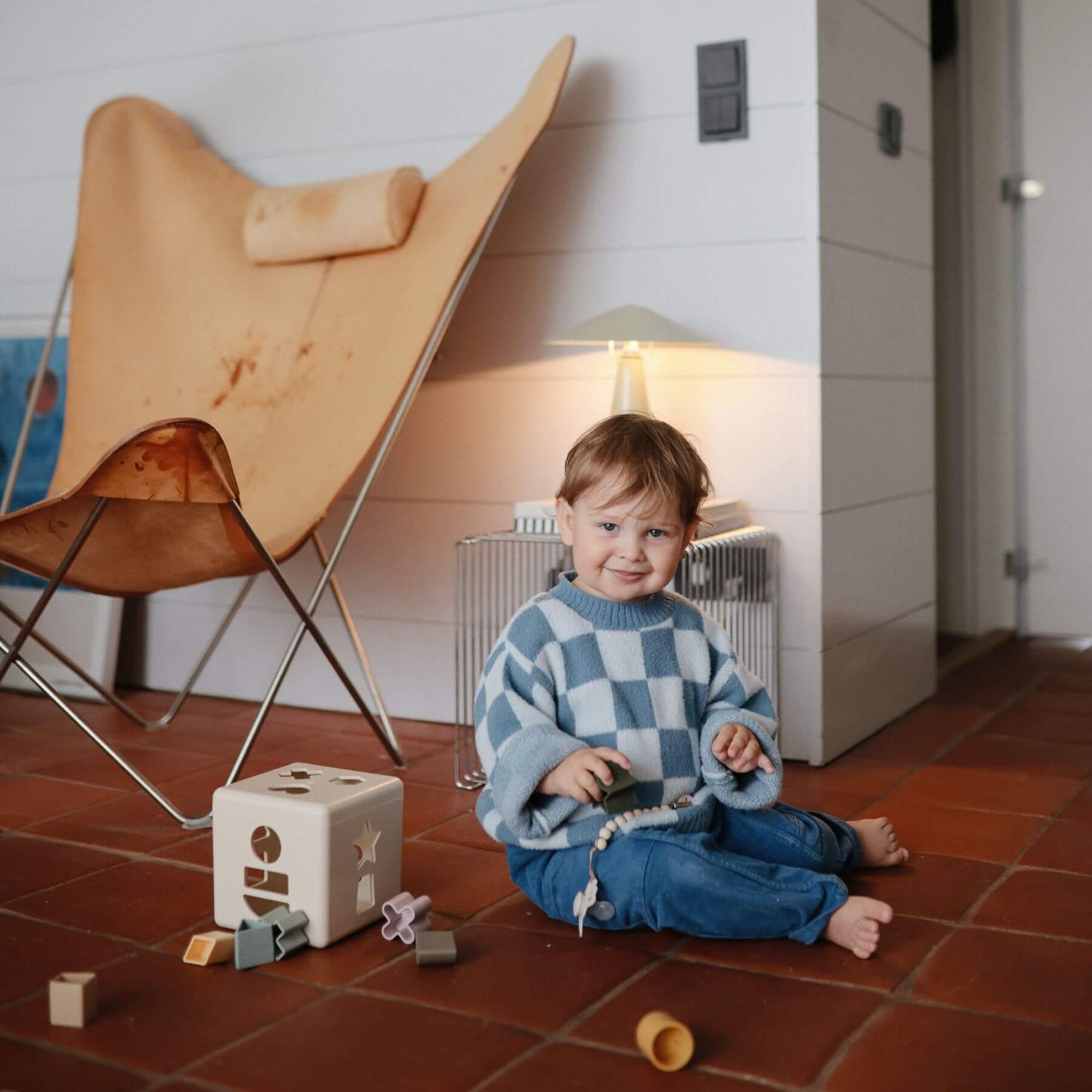 Child sitting on a tiled floor with Mushie Shape Sorting Box, wearing a blue checkered sweater and jeans, in a room with a wooden chair and lamp.