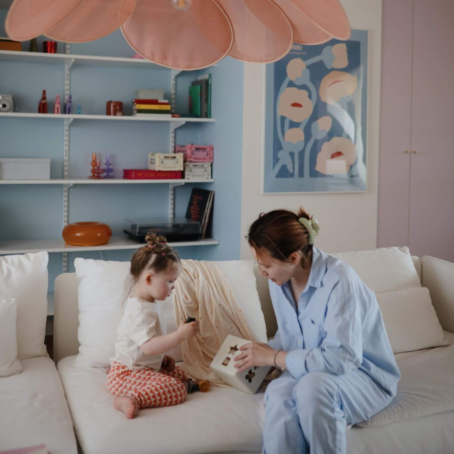 Woman and child sitting on a couch with Mushie Shape Sorting Box, in a room with shelves and artwork.