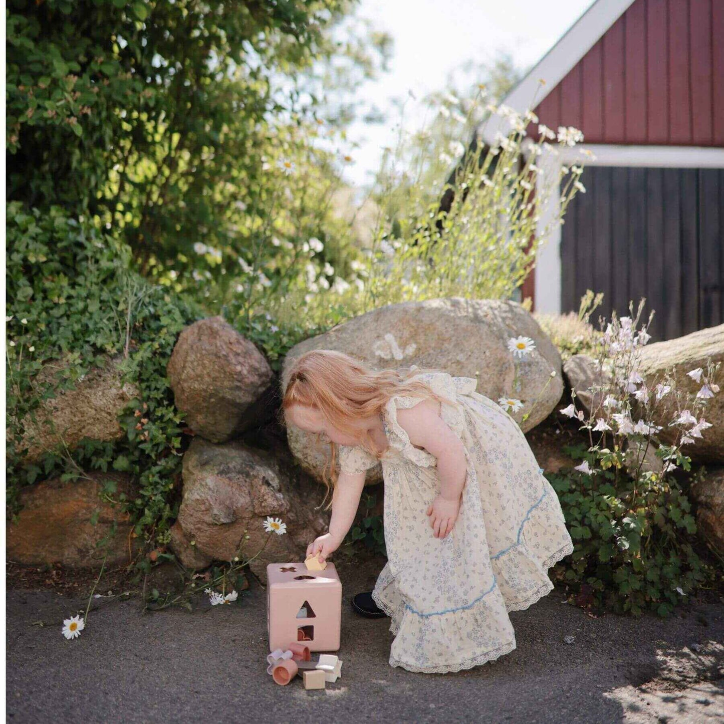 Child in a white dress playing with Mushie Shape Sorting Box outdoors near rocks and flowers.