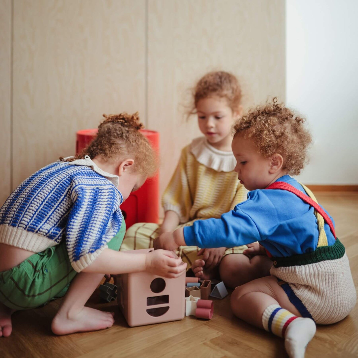 Three children playing with Mushie Shape Sorting Box on a wooden floor.