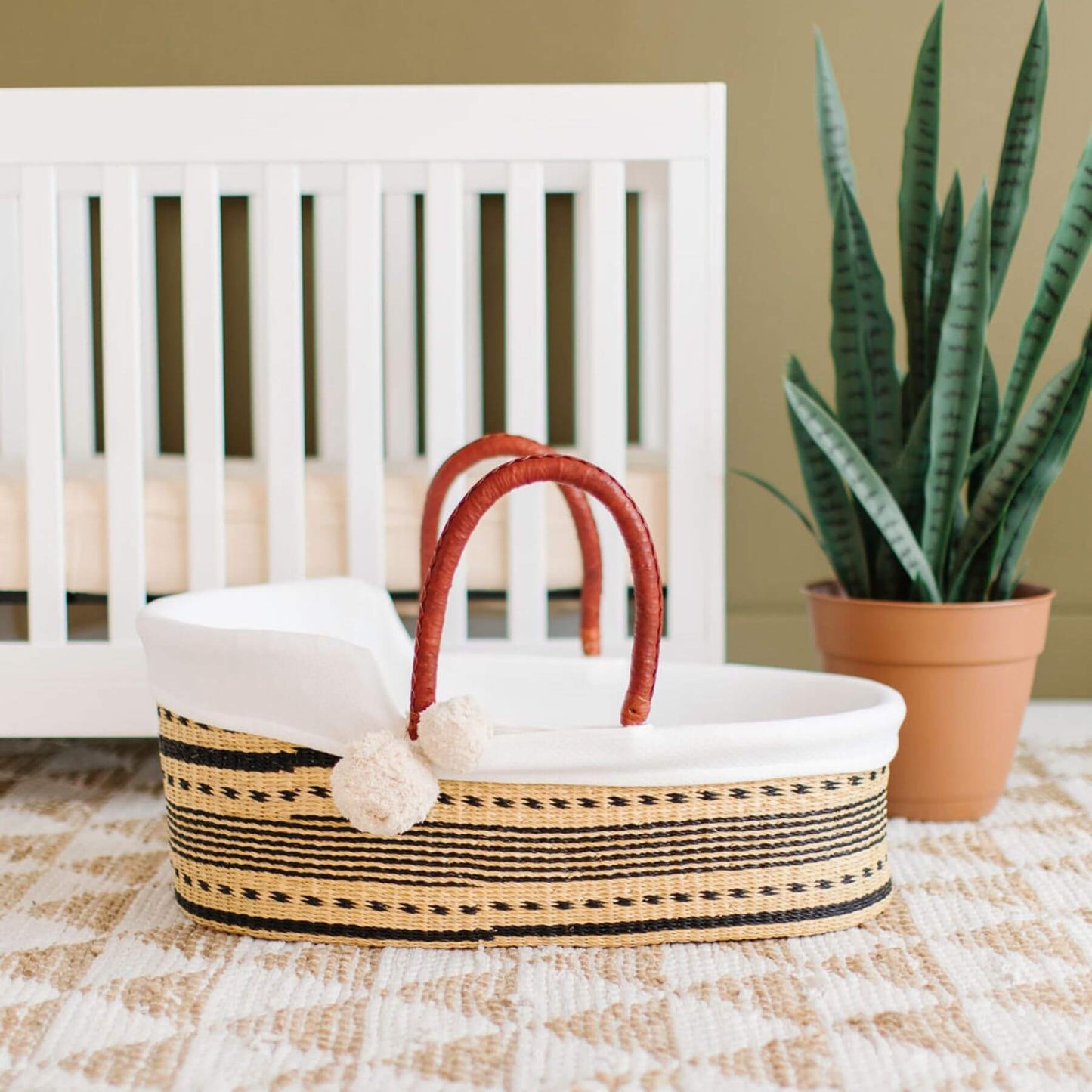 Spring Sparrow African Moses Basket in front of a white crib with a plant in the background.