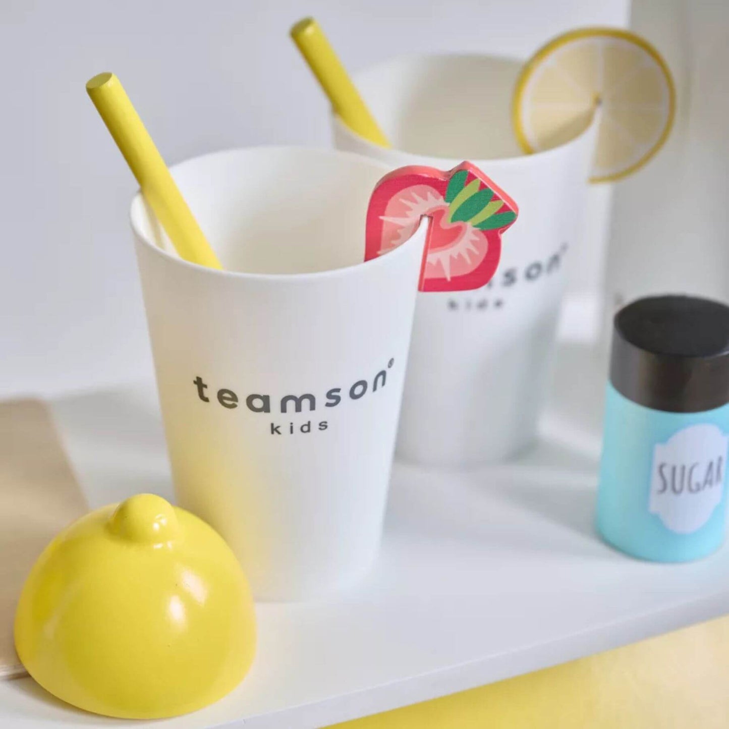 White cup with yellow straw, lemon-shaped container, and 'teamson kids' packaging on a white surface.